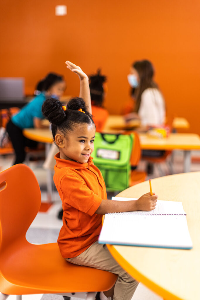 Girl in orange shirt raising hand in classroom setting