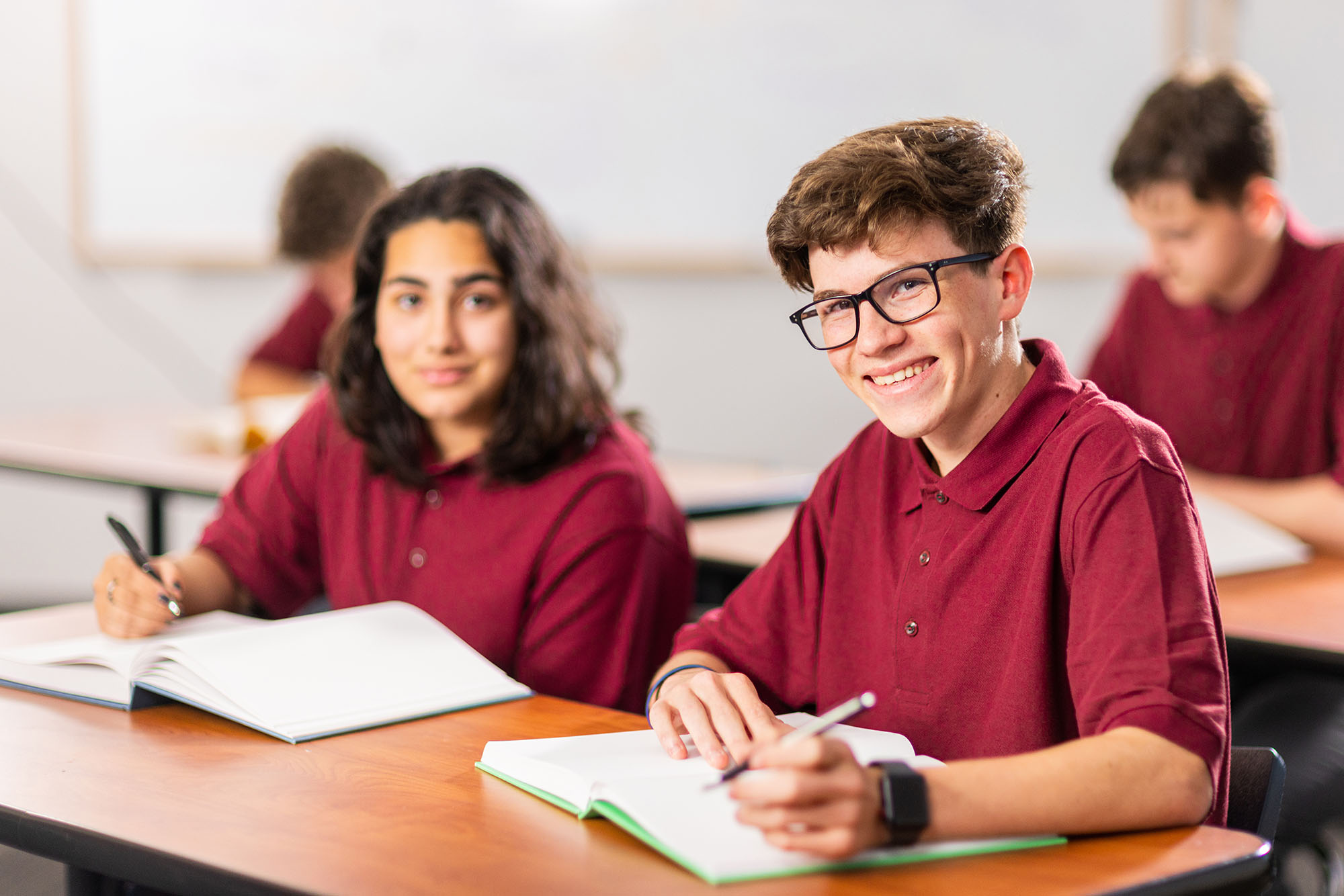 Students in maroon shirts smiling at school desk